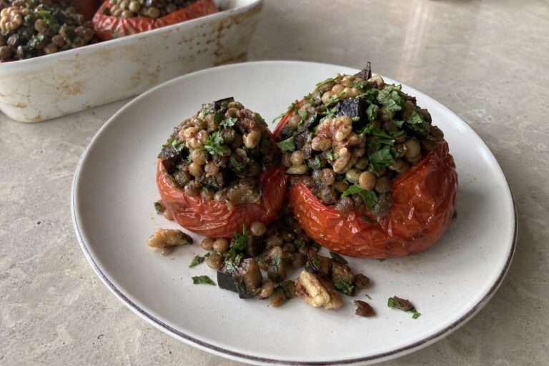 Two baked stuffed tomatoes with wrinkled red skins on a plate, filled with dark brown eggplant cubes, brown lentils, toasted walnut pieces and chopped parsley and mint, with a baking dish in the background.