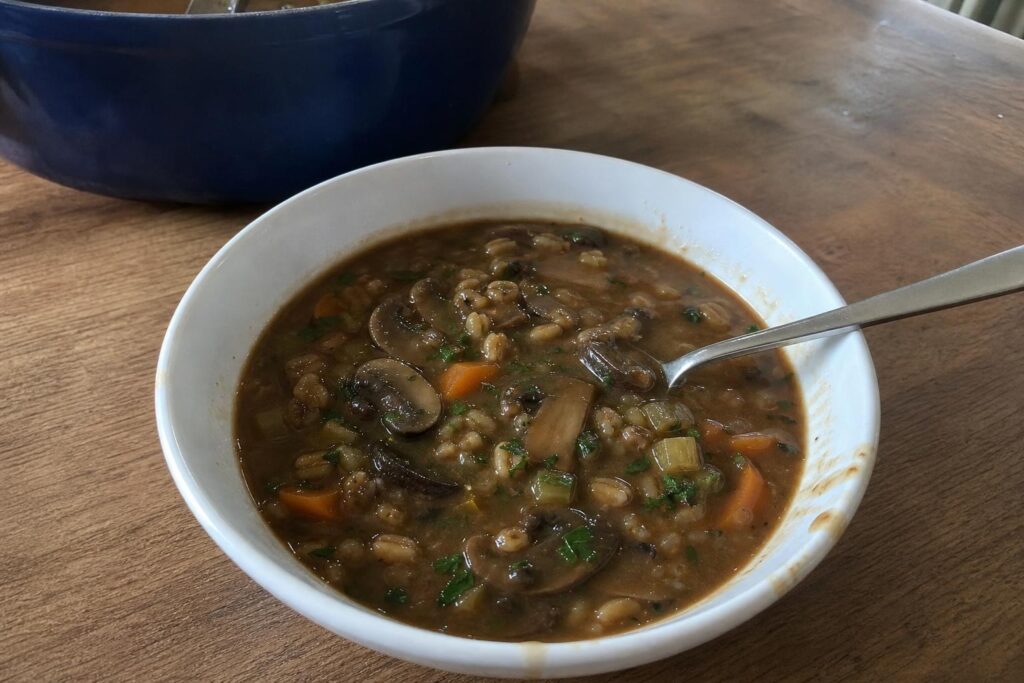 A deep everyday bowl of mushroom barley soup with a dark thick broth, sliced cremini and shiitake mushrooms, carrot half moons, celery pieces, swollen barley grains and a scattering of chopped parsley, with a spoon resting in the bowl and the pot in the background.