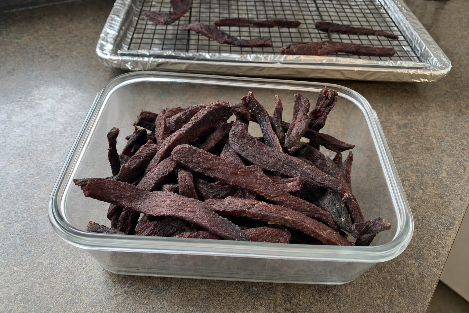 Homemade AIP beef jerky strips in a glass container, with the drying rack behind it