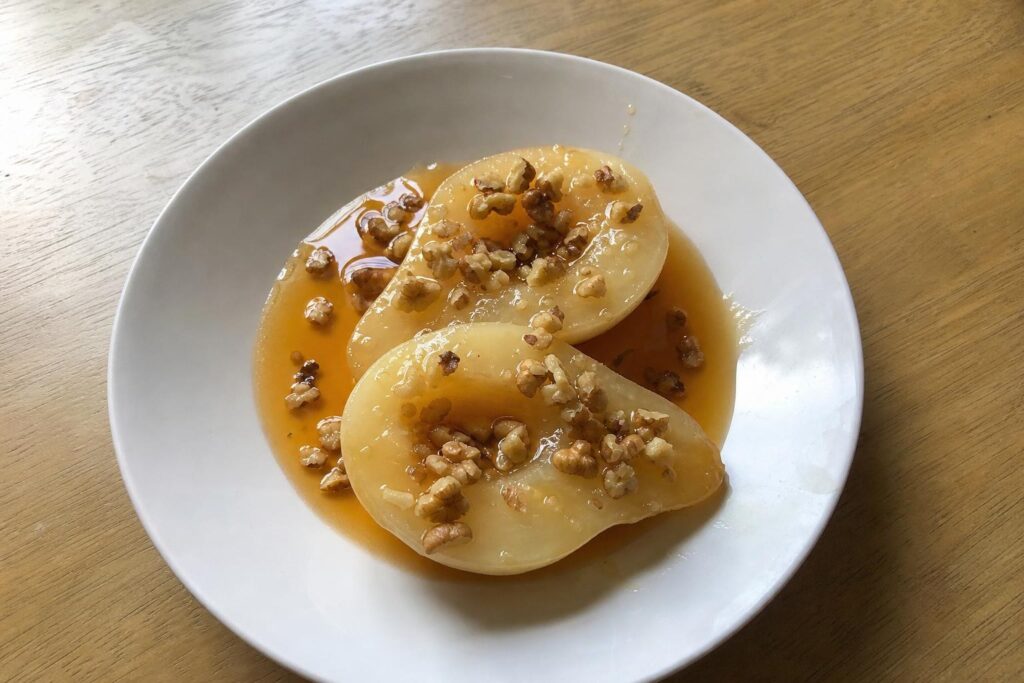 Two peeled pear halves in a shallow white bowl, core side up, glazed with a thin orange syrup and topped with coarsely chopped toasted walnuts.