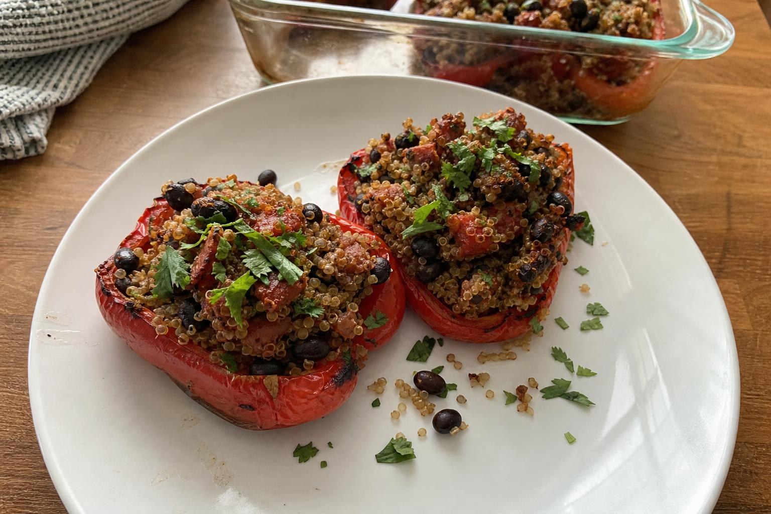 Two red bell pepper halves filled with quinoa, whole black beans and diced tomato on a plain dinner plate, topped with chopped cilantro, with a baking dish out of focus behind them.