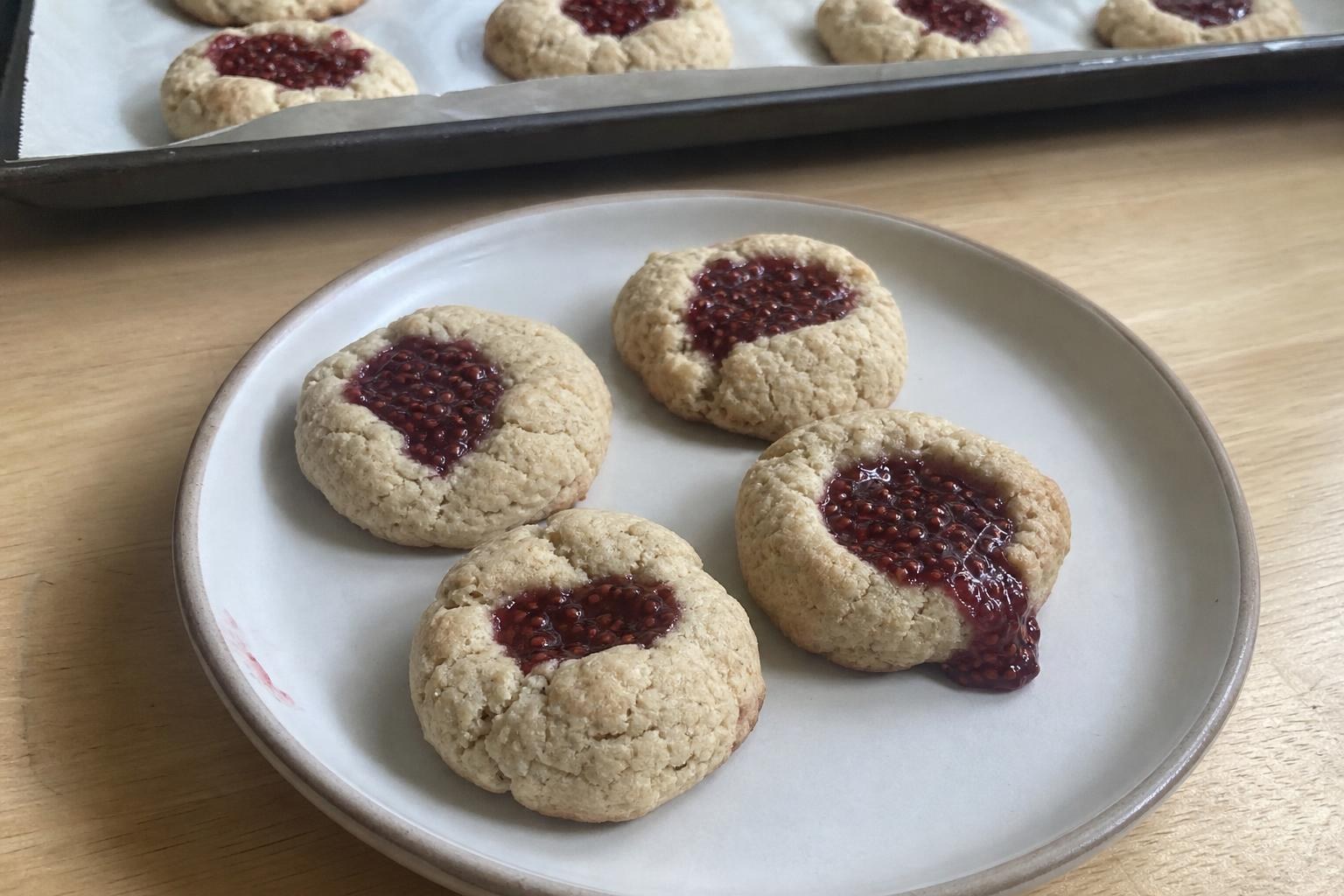 A small everyday plate with four homemade raspberry chia jam thumbprint cookies, pale beige with golden edges and matte, slightly cracked tops, each hand-pressed well filled with dark red chia jam, with a parchment-lined baking sheet holding more cookies blurred behind.