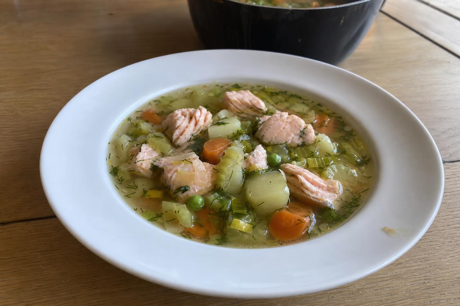 A deep everyday bowl of pale golden clear salmon and vegetable soup with large pale pink poached salmon cubes, potato chunks, carrot coins, soft leek half-moons, green peas and chopped fresh dill on top.