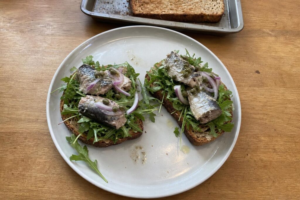 Two open-faced toasts of whole-grain bread topped with baby arugula, large irregular pieces of canned sardine, thin slices of red onion and chopped capers, glistening with lemon olive oil dressing on a plain plate.