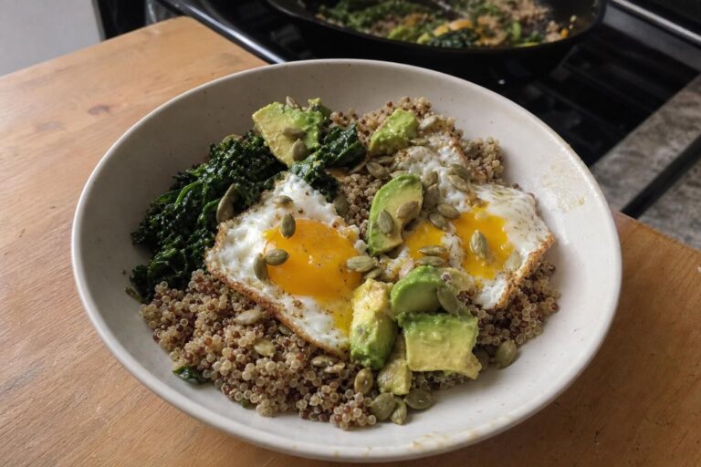 A shallow ceramic bowl of savory breakfast food on a kitchen table, filled with loose cooked quinoa, dark green sautéed kale piled to one side, two fried eggs with set whites and golden lacy edges and runny yellow yolks on top, uneven hand-sliced avocado and toasted pumpkin seeds scattered over the bowl, with a blurred skillet in the background.