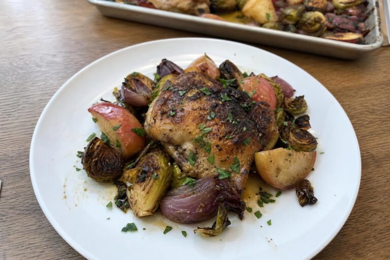 Una ración individual de pollo al horno con coles de Bruselas y manzanas en un plato, en la que se ve un muslo de pollo asado, coles de Bruselas cortadas por la mitad y doradas, gajos de manzana tiernos con piel, gajos de cebolla roja y perejil picado.