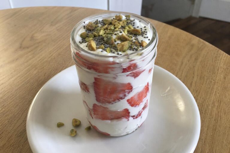 A glass jar parfait with layers of thick white Greek yogurt and sliced fresh strawberries, topped with chopped toasted pistachios and chia seeds.
