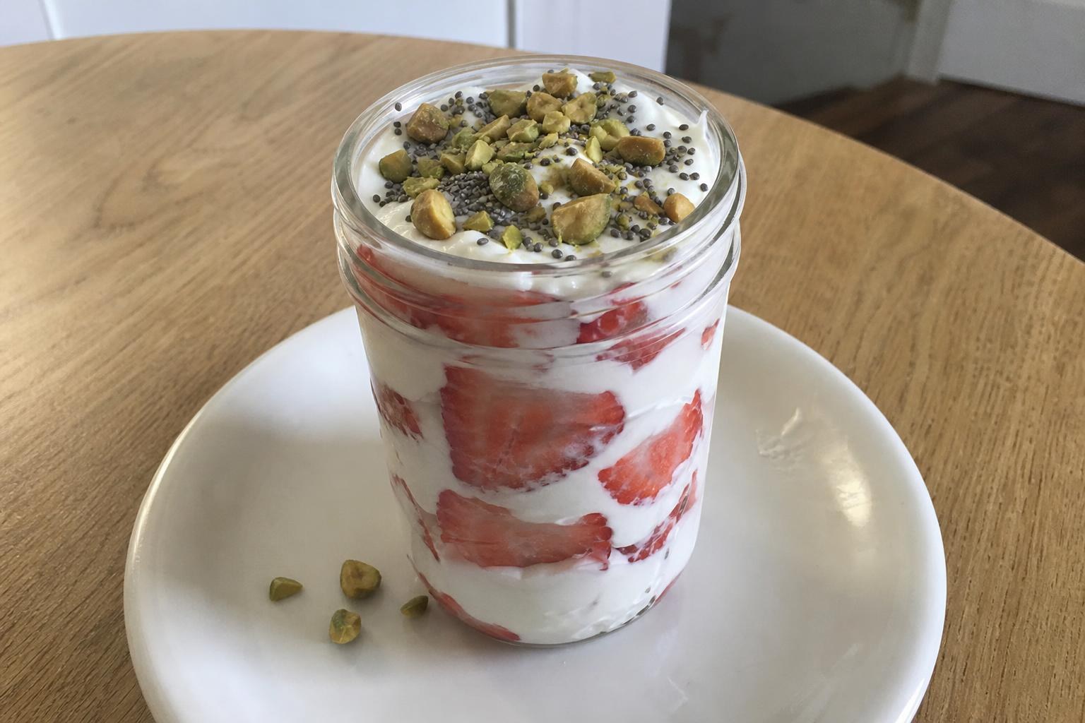 Un parfait en un tarro de cristal con capas de yogur griego blanco espeso y fresas frescas cortadas en rodajas, coronado con pistachos tostados picados y semillas de chía.