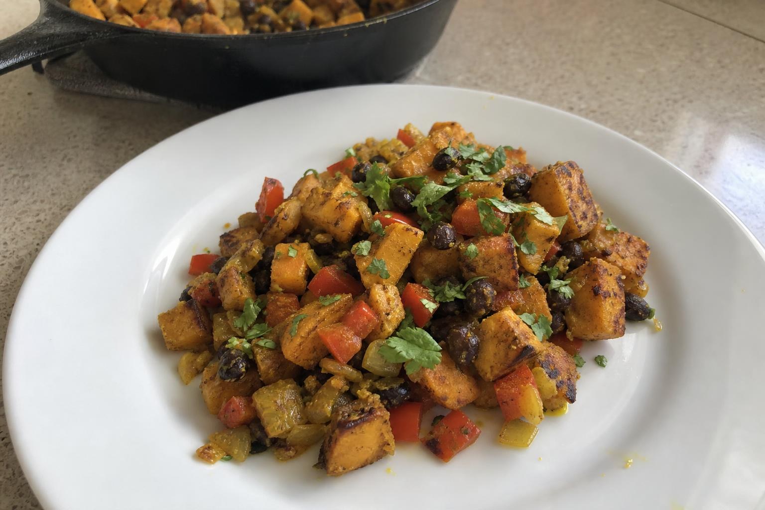 Una ración de revuelto de boniato y alubias negras para desayunar en un plato sencillo: cubitos dorados de boniato, alubias negras enteras, trozos de pimiento rojo y cebolla, teñidos de un tono amarillo anaranjado por la cúrcuma y coronados con cilantro picado, con una sartén de hierro fundido difuminada al fondo.