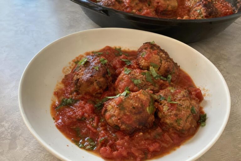 A shallow bowl of turkey and zucchini meatballs with tomato sauce, hand-rolled and uneven in size, nestled in a chunky crushed tomato sauce and topped with chopped fresh basil.