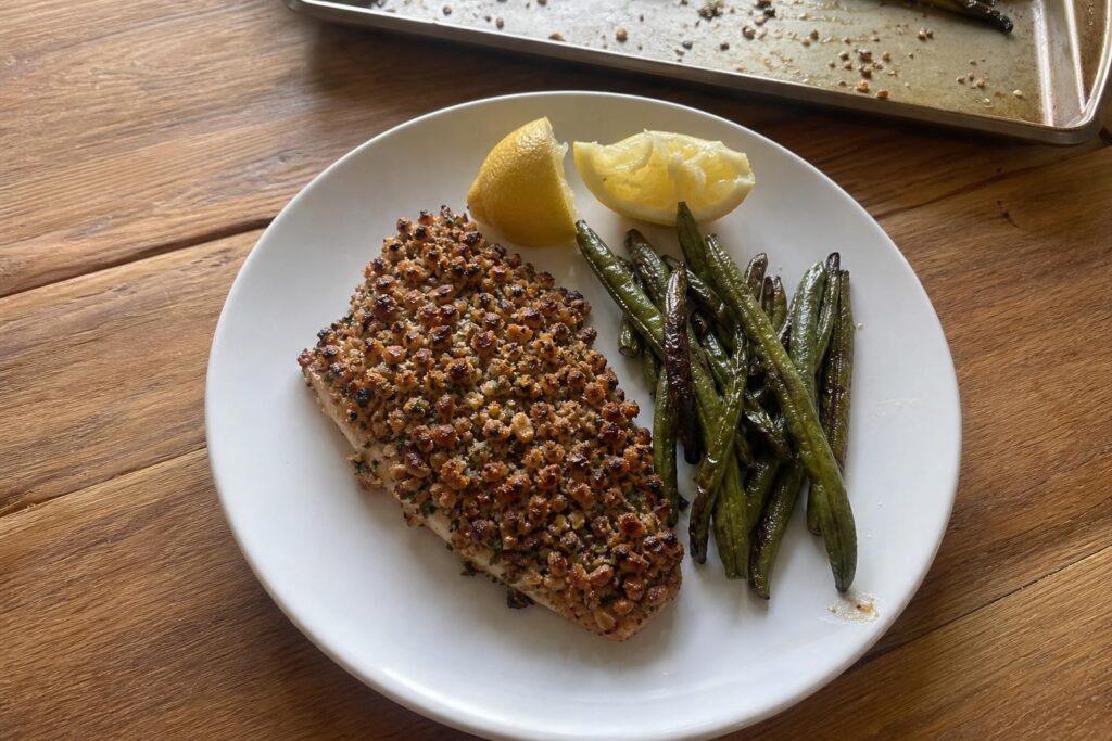 A fillet of walnut-crusted trout with a craggy golden brown chopped walnut topping, skin side down on a white plate next to roasted green beans and two lemon wedges.