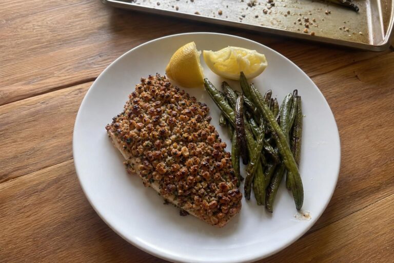 A fillet of walnut-crusted trout with a craggy golden brown chopped walnut topping, skin side down on a white plate next to roasted green beans and two lemon wedges.