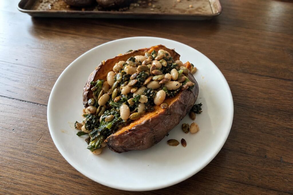 A dinner plate with one whole baked sweet potato split lengthwise and open, its fluffed orange flesh filled with white beans, dark green wilted kale and toasted pumpkin seeds, with a baking sheet in the background.